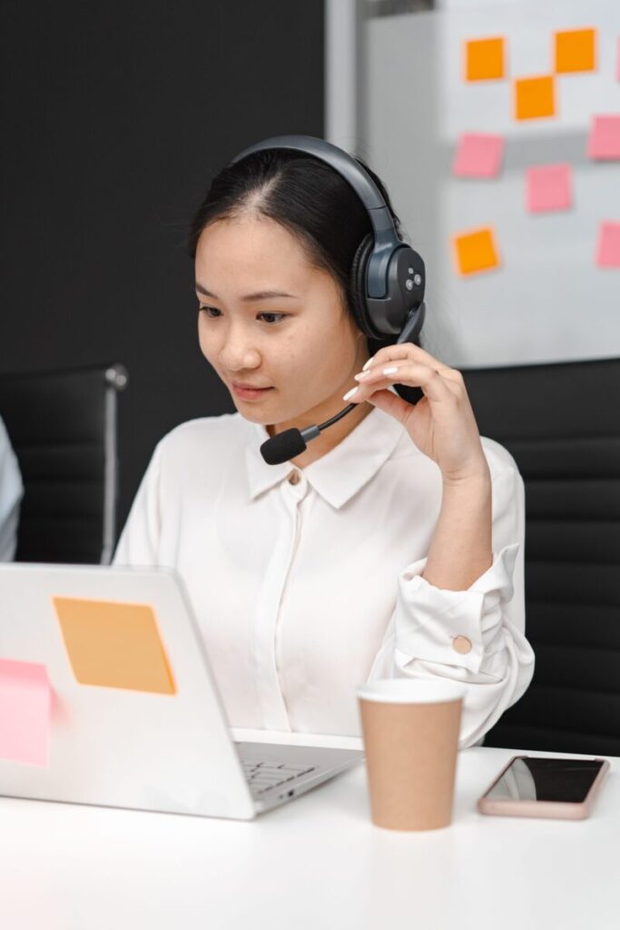 Asian woman using a laptop and headset for customer support in an office environment.